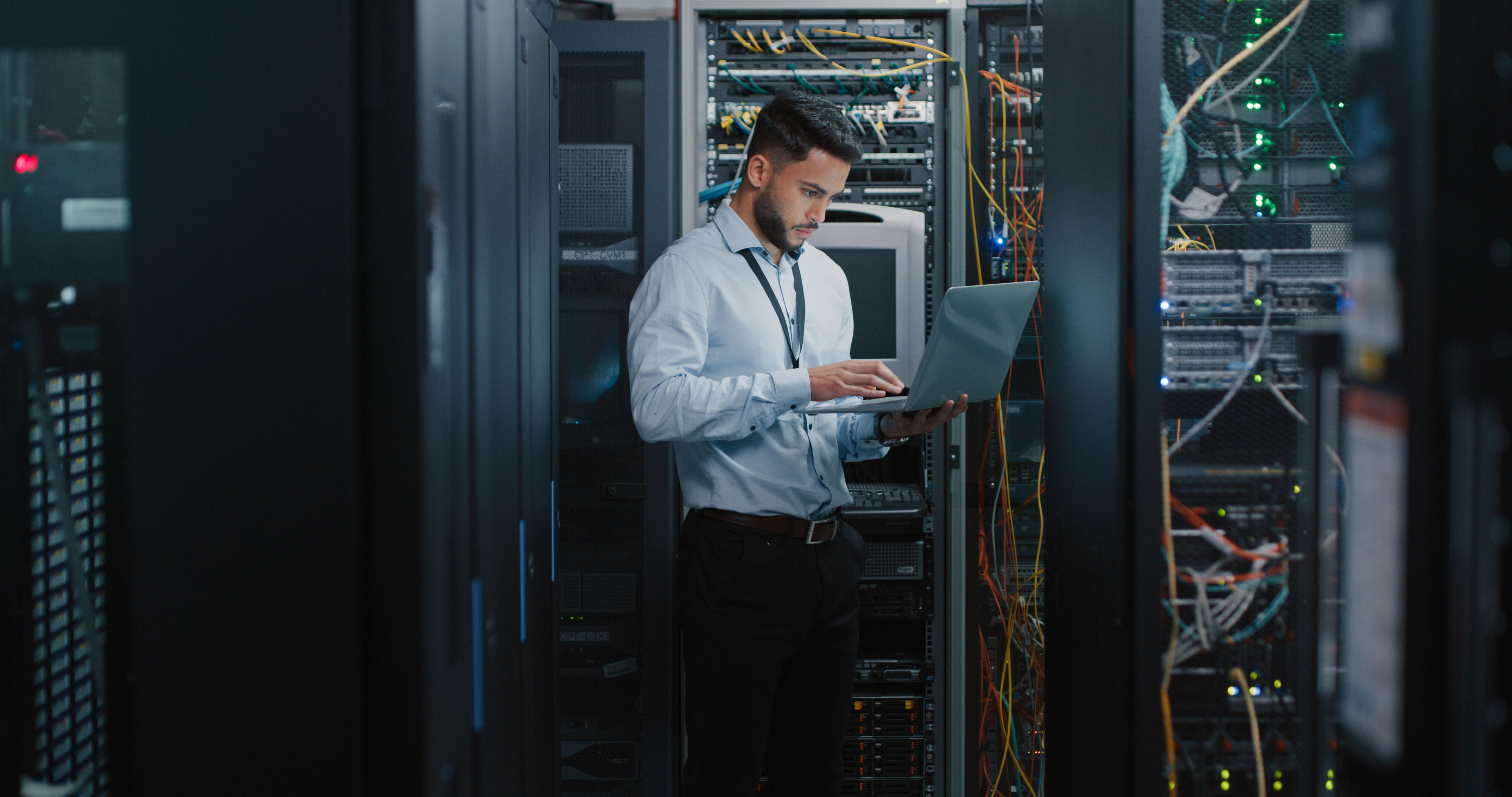 A man standing among network servers while looking down intently at a laptop