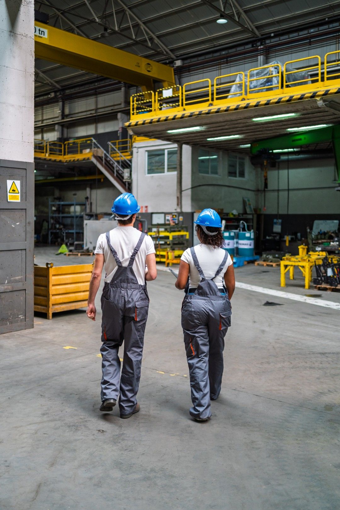 Two workers in hard hats and overalls walking into a workshop bay beneath an overhead crane
