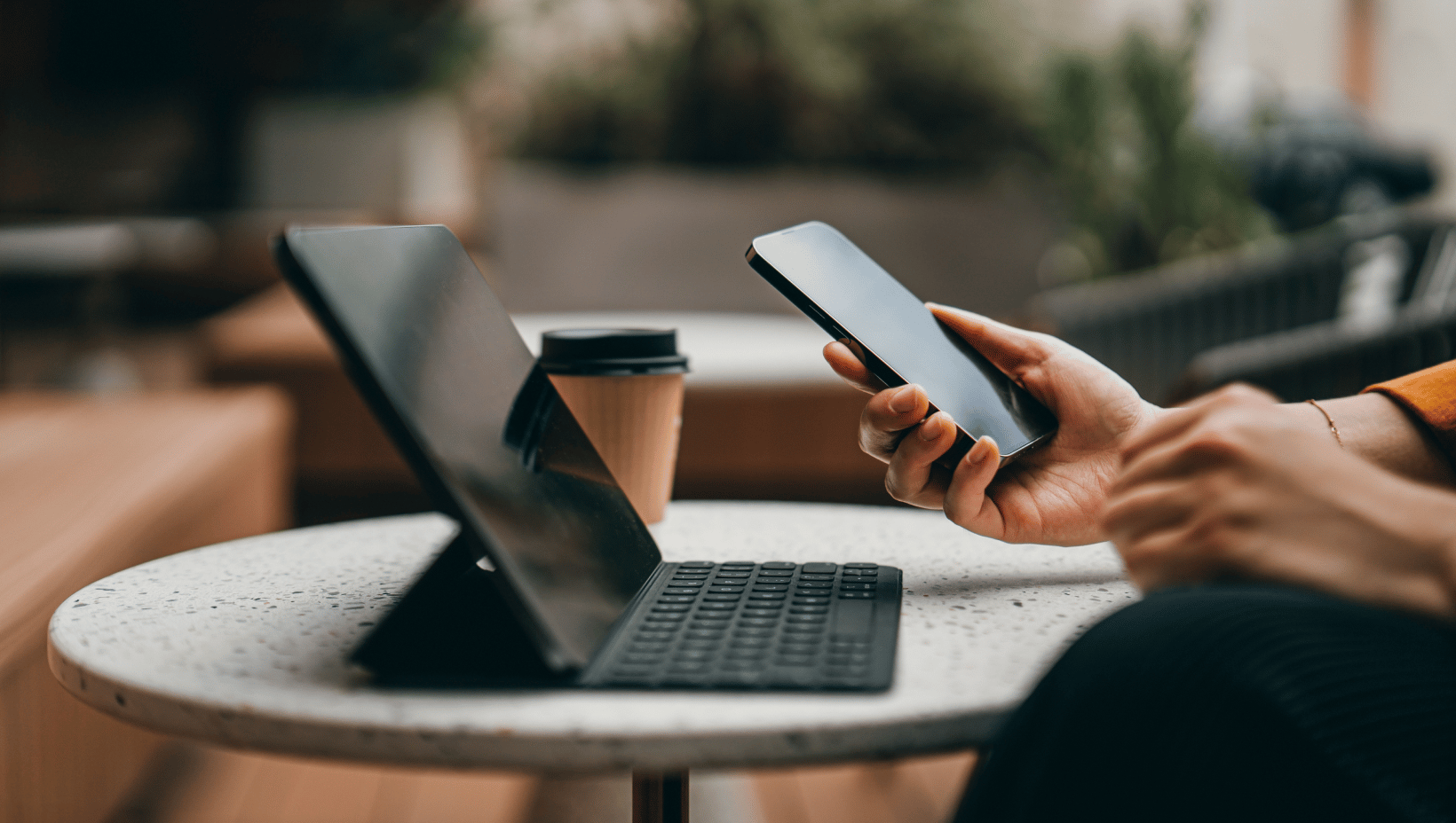 Close-up of a professional holding a smartphone beside a tablet with a keyboard and a coffee cup on a café table