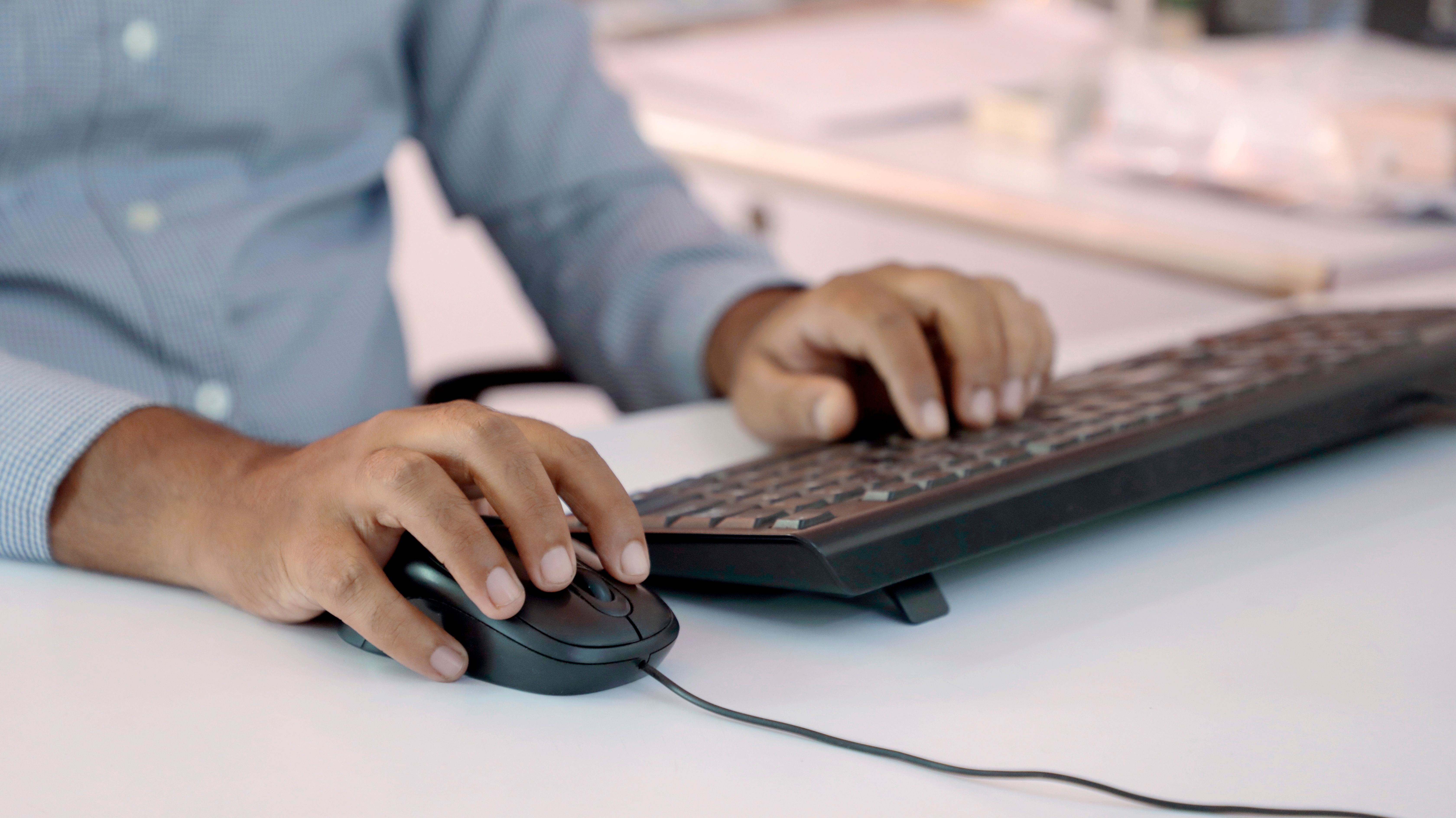 Close-up of a man's hands in a blue shirt using a keyboard and corded mouse at a white office desk