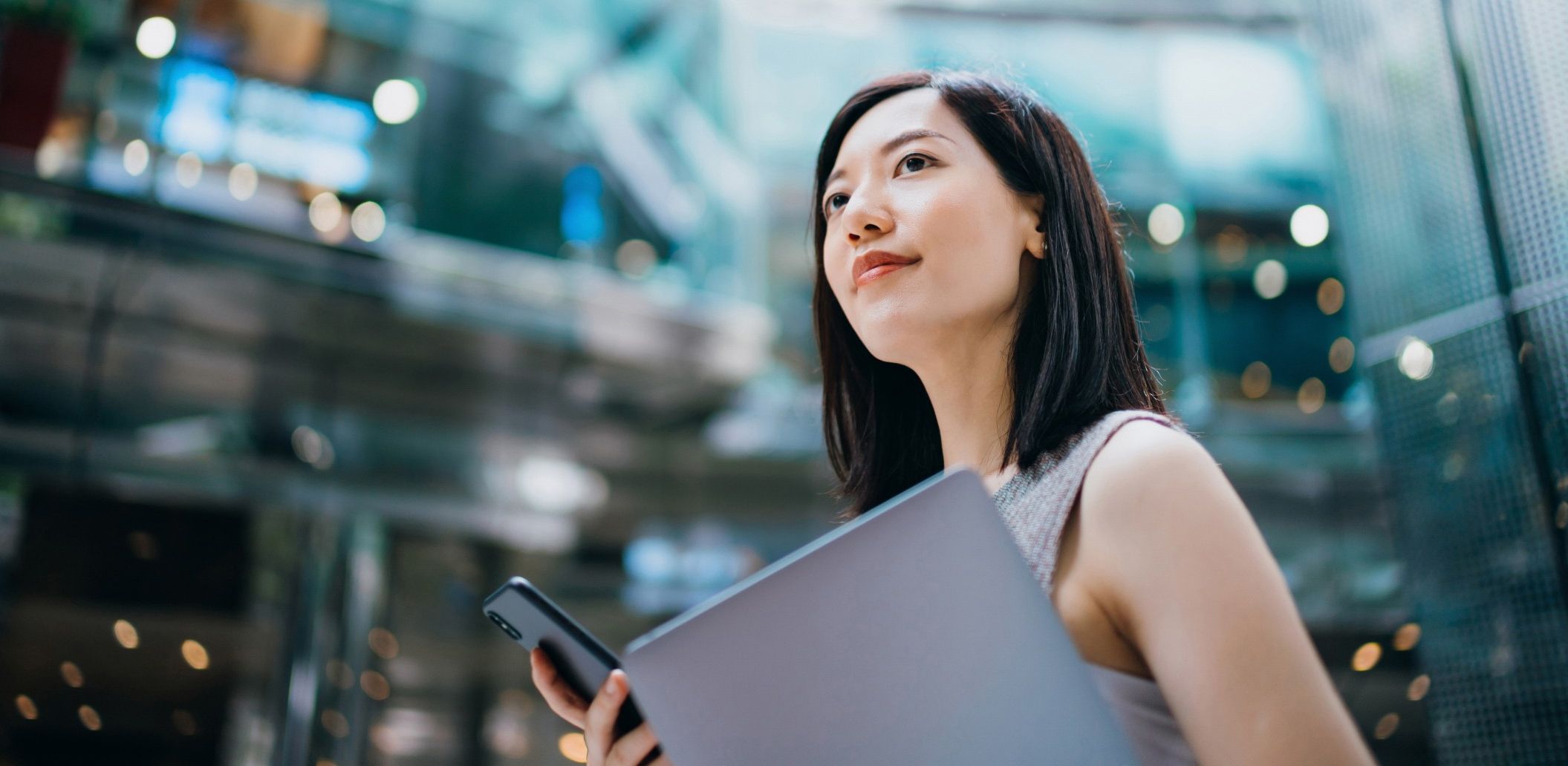 Woman with shoulder-length dark hair carrying a company laptop and smartphone in a modern glass office building