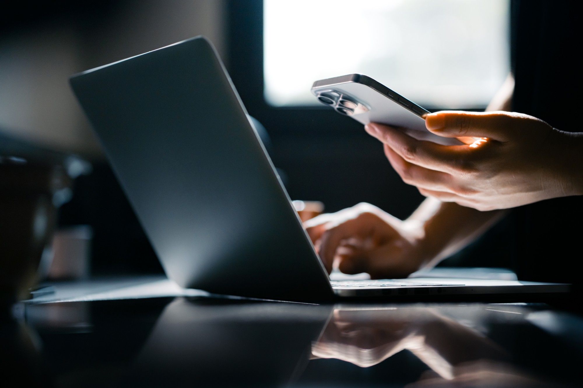 Close-up of a professional's hands typing on a business laptop while holding a smartphone at a dark desk