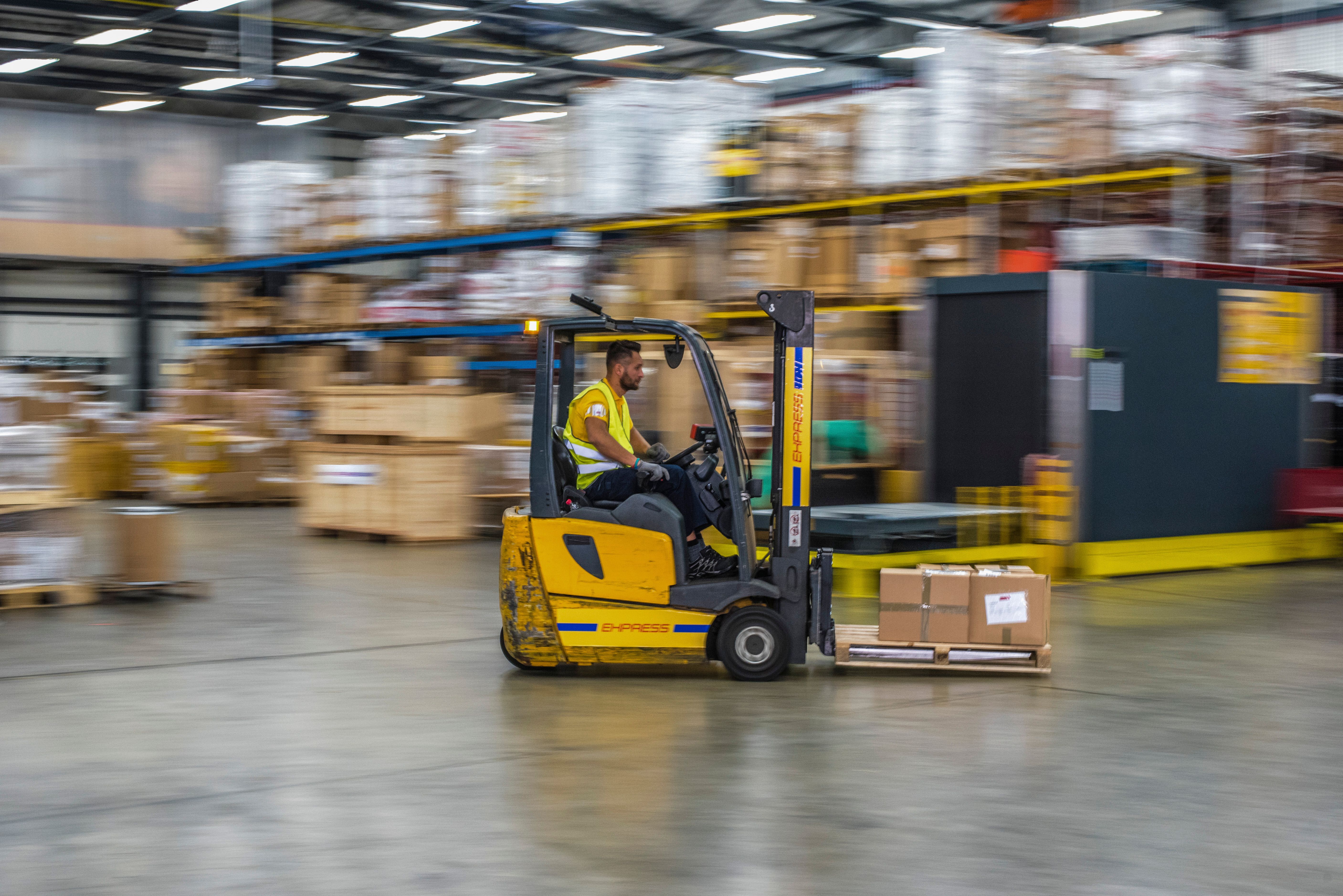 Operator driving a yellow counterbalance forklift carrying a pallet across a warehouse floor