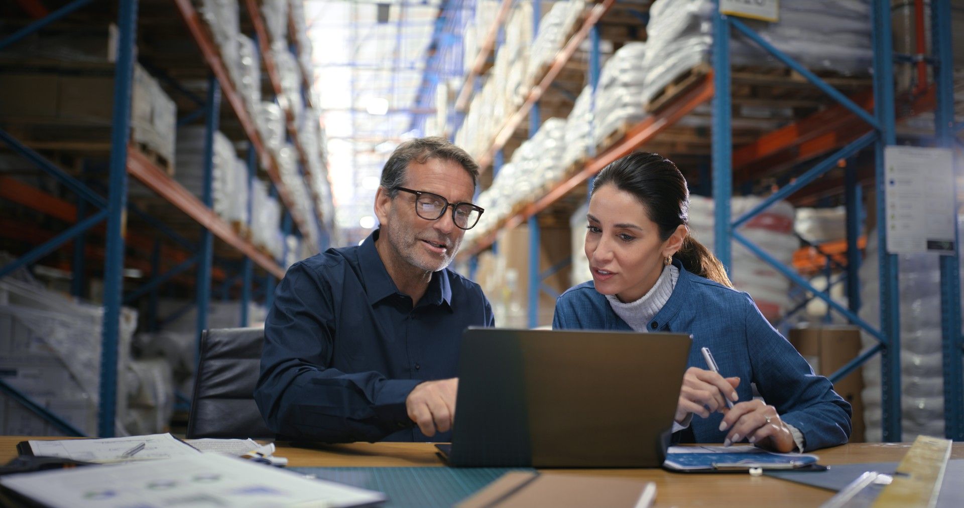Two colleagues reviewing a laptop together in a warehouse aisle lined with wrapped pallets