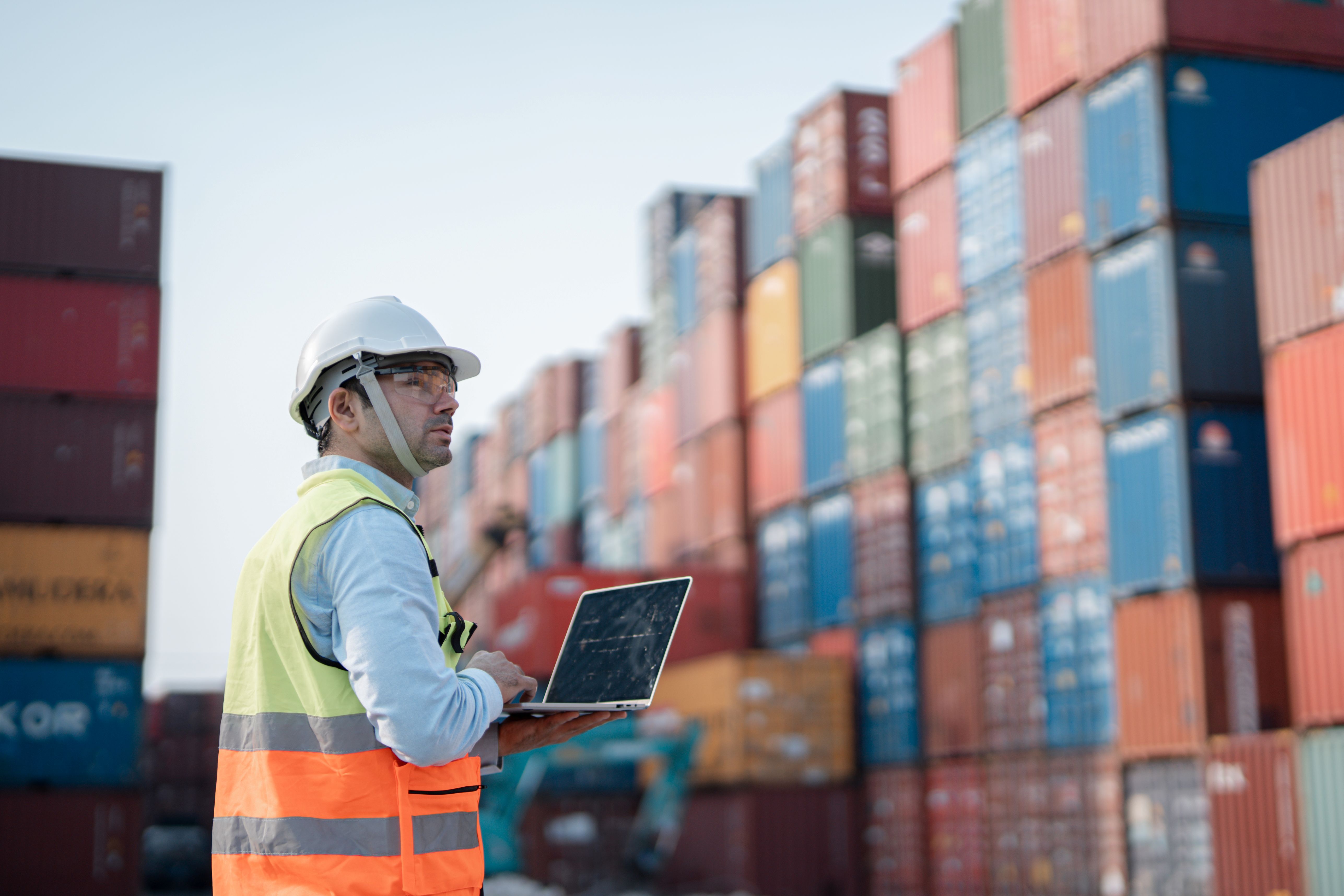 Logistics worker in a hard hat and high-visibility vest using a laptop at a container yard, with stacked shipping containers behind him.
