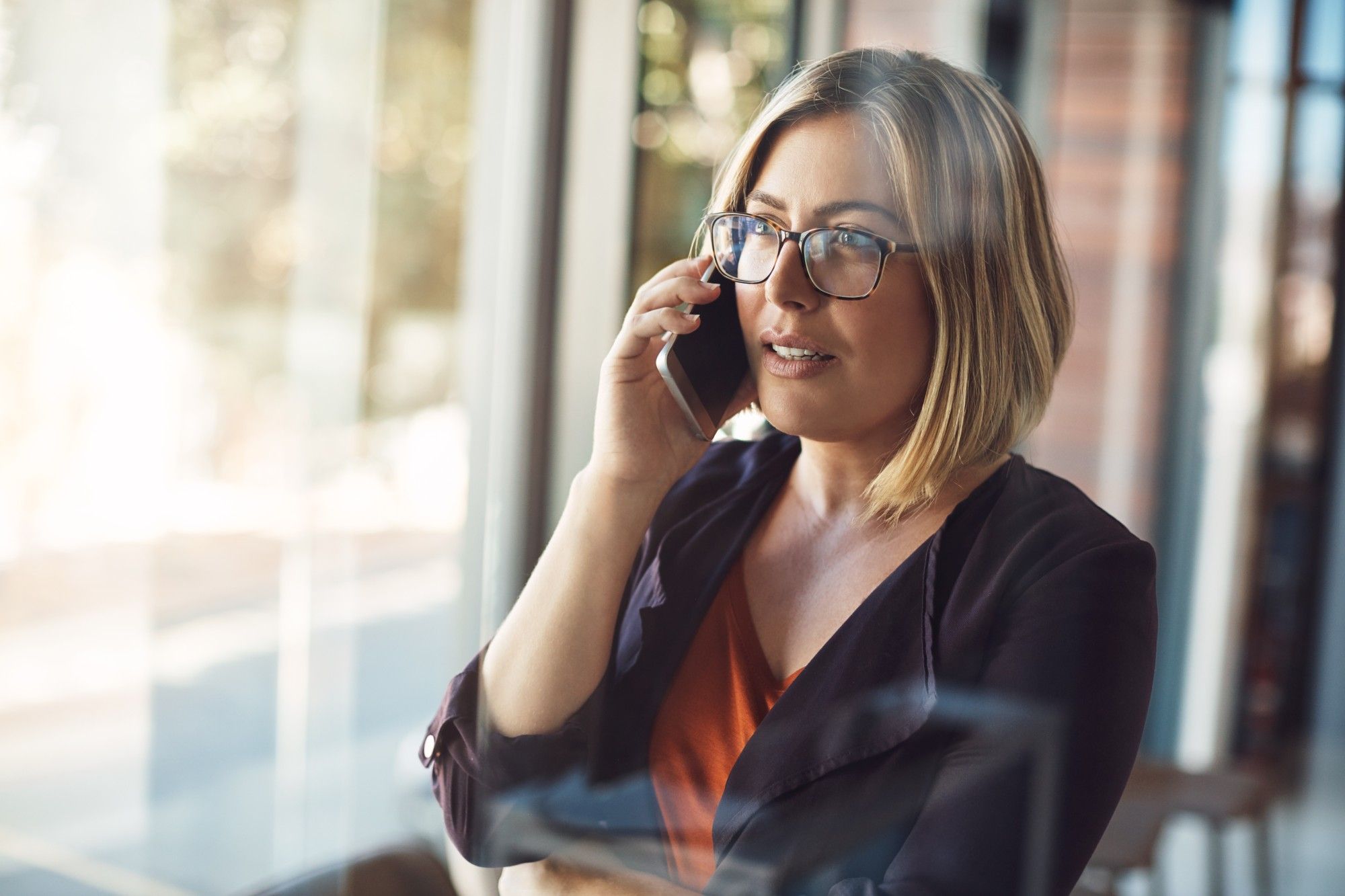 Woman with blonde hair and glasses in a black blazer talking on her smartphone beside an office window