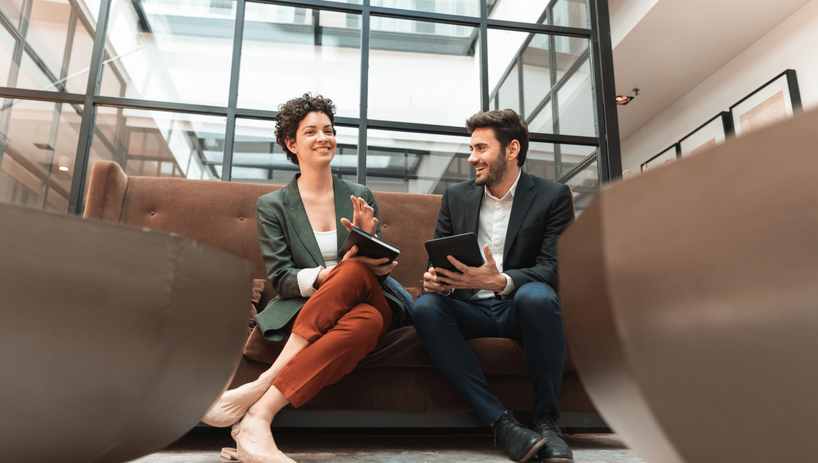 Smiling businesswoman in a green blazer and businessman in a dark suit discussing plans on tablets in a modern office lobby