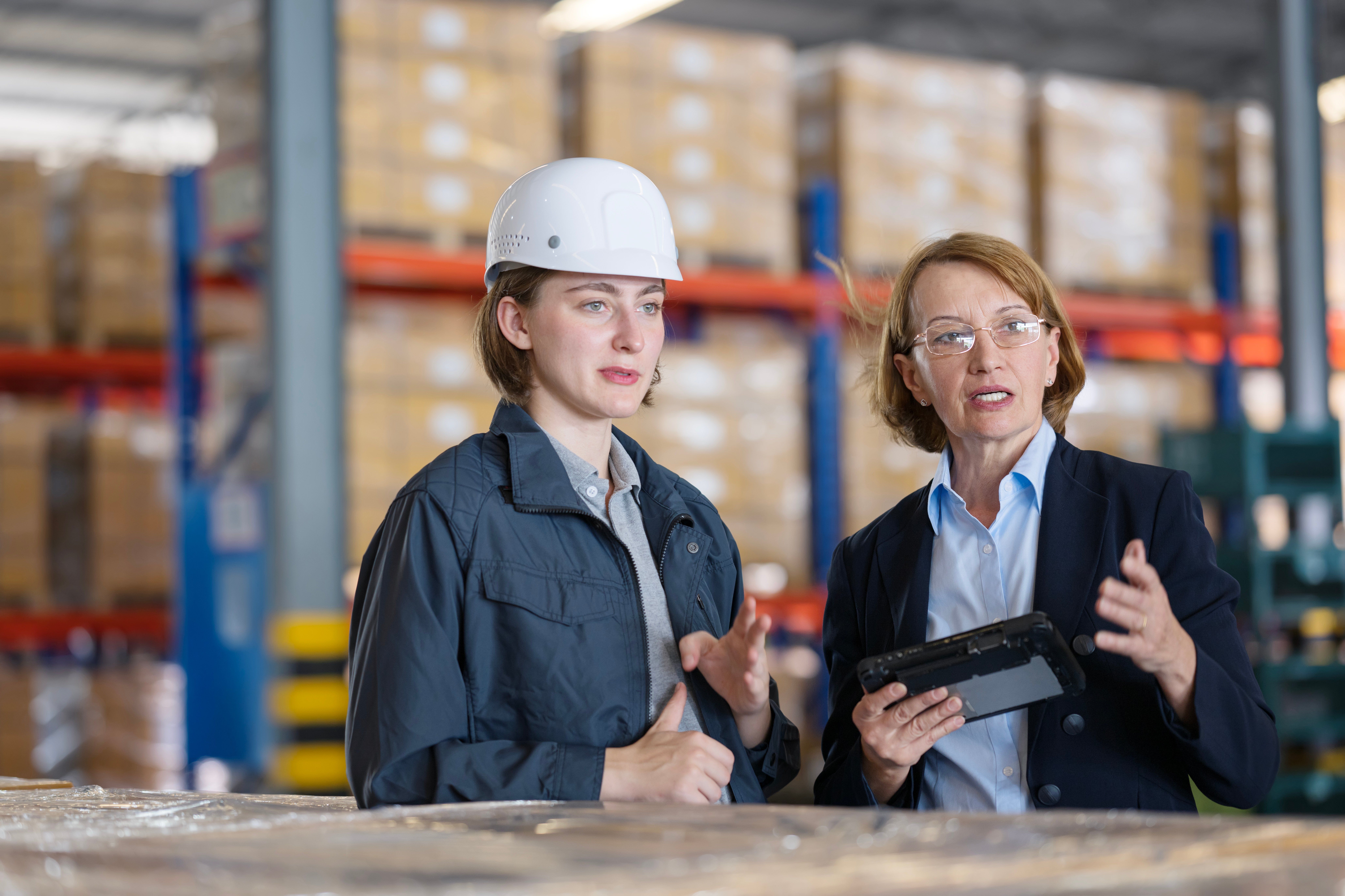 Two woman in a warehouse talking. One is in a business suite and another in warehouse attire