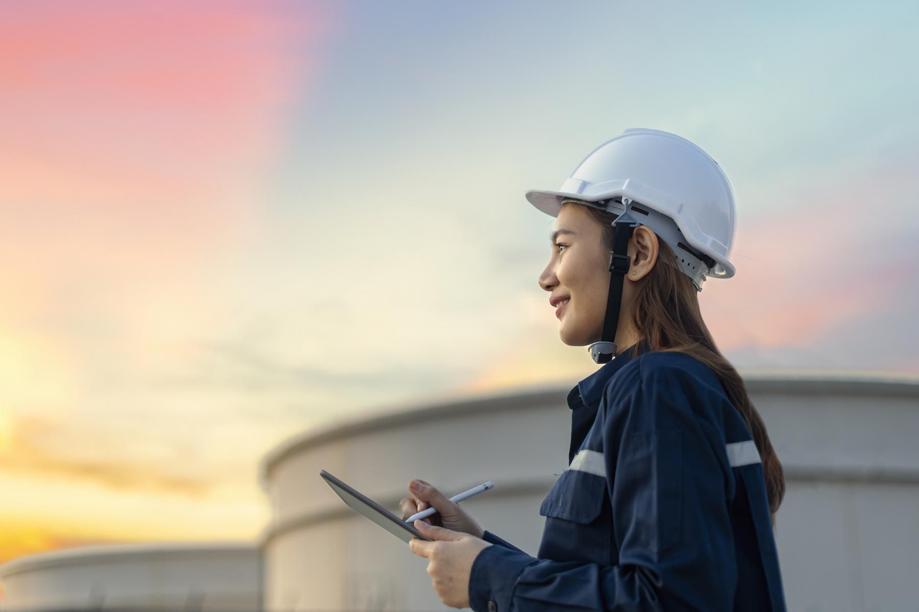 A worker in a hard hat and coveralls holding a tablet, inspecting large industrial storage tanks at sunset.