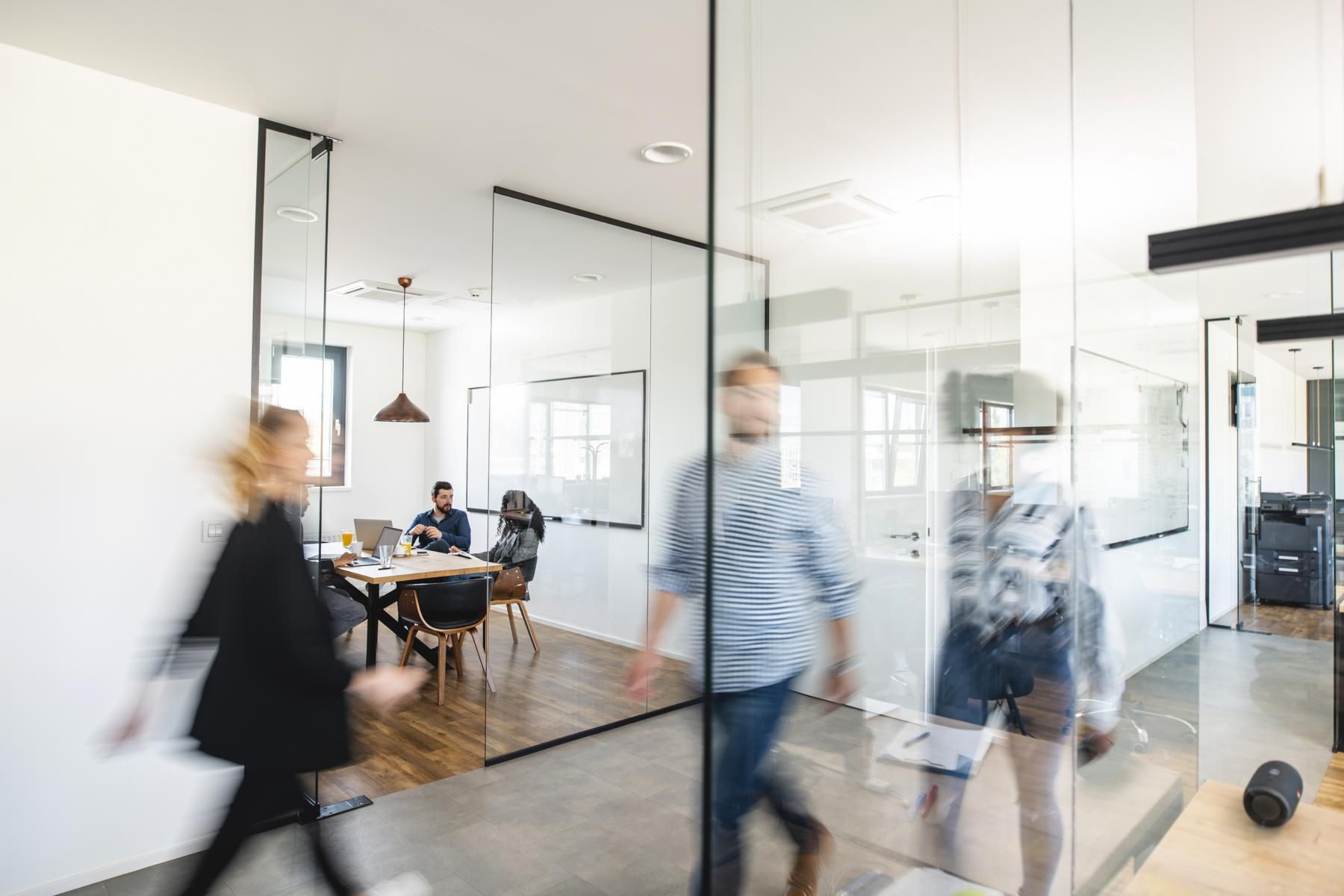 A team meeting in progress inside a glass-walled office as colleagues walk past.