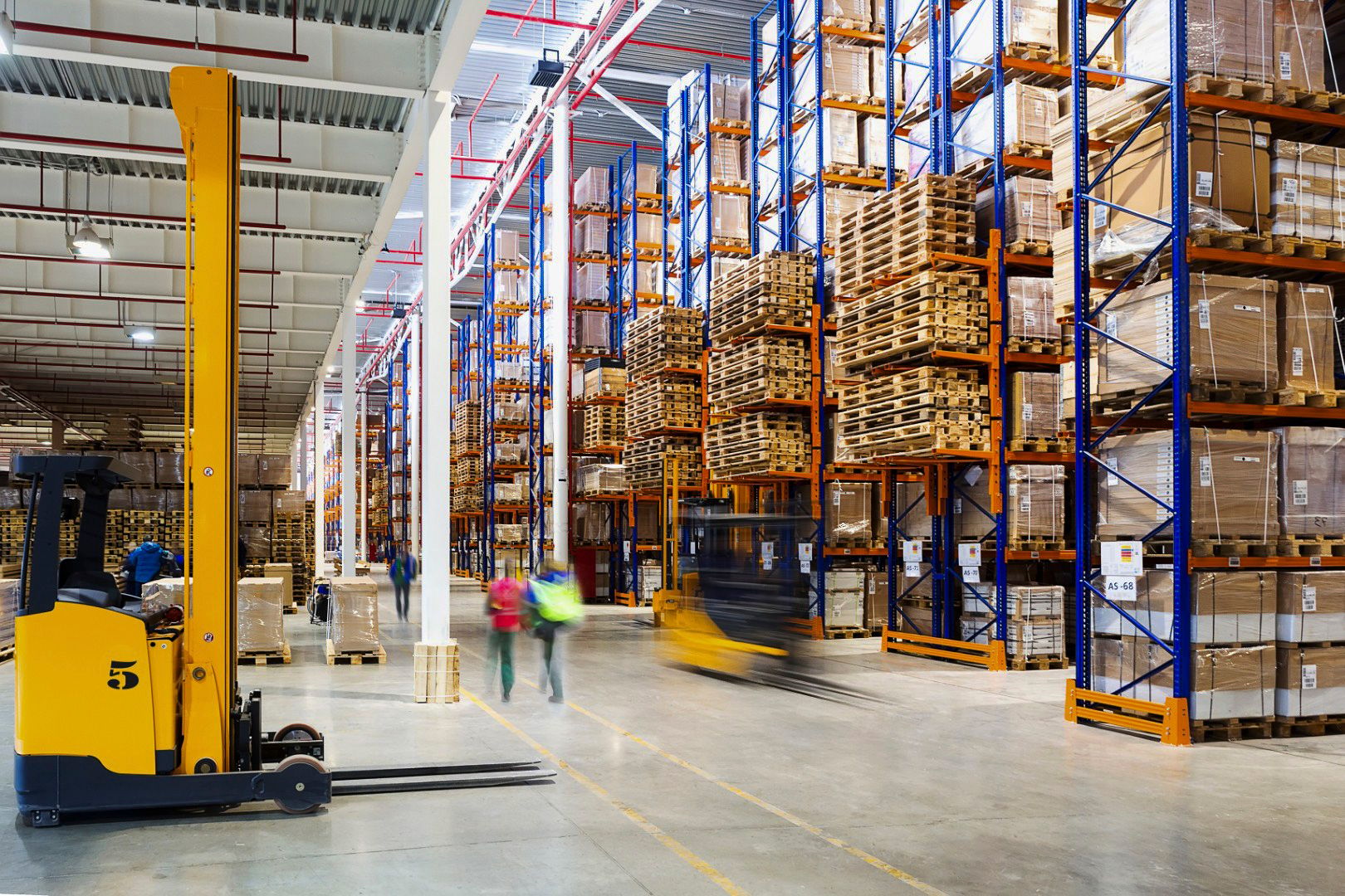 High-bay warehouse with pallets stacked on orange racking as forklifts and workers move through the aisle