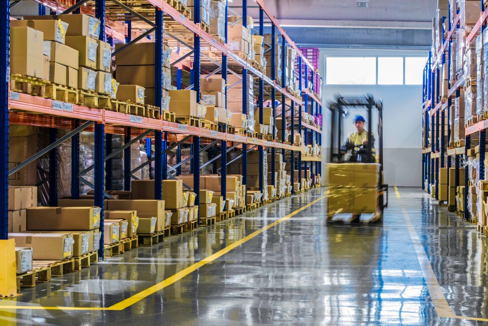 Operator on a pallet truck at the far end of a warehouse aisle lined with boxes on blue pallet racking
