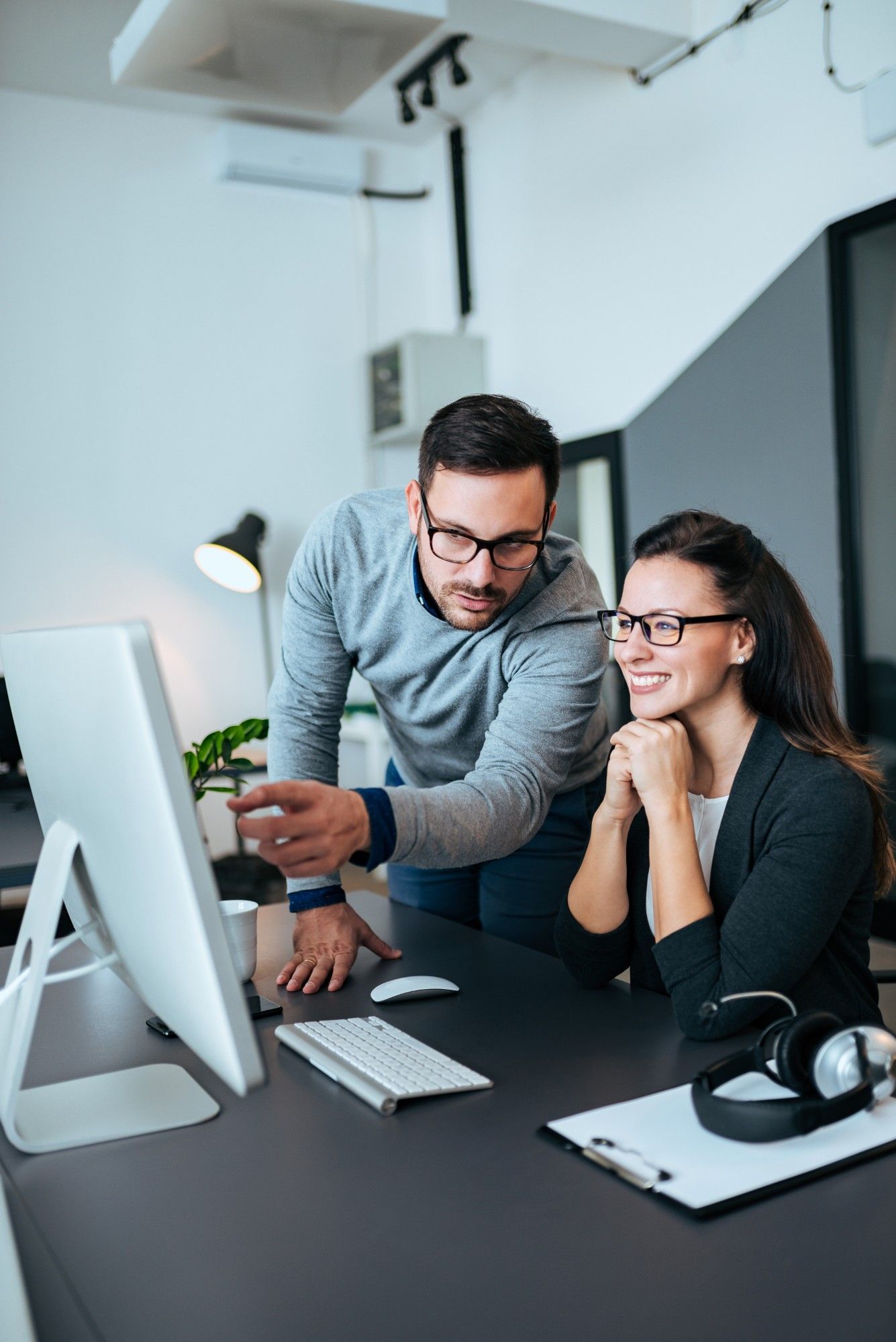 Man in a gray sweater pointing at an all-in-one desktop computer screen as a smiling female colleague looks on