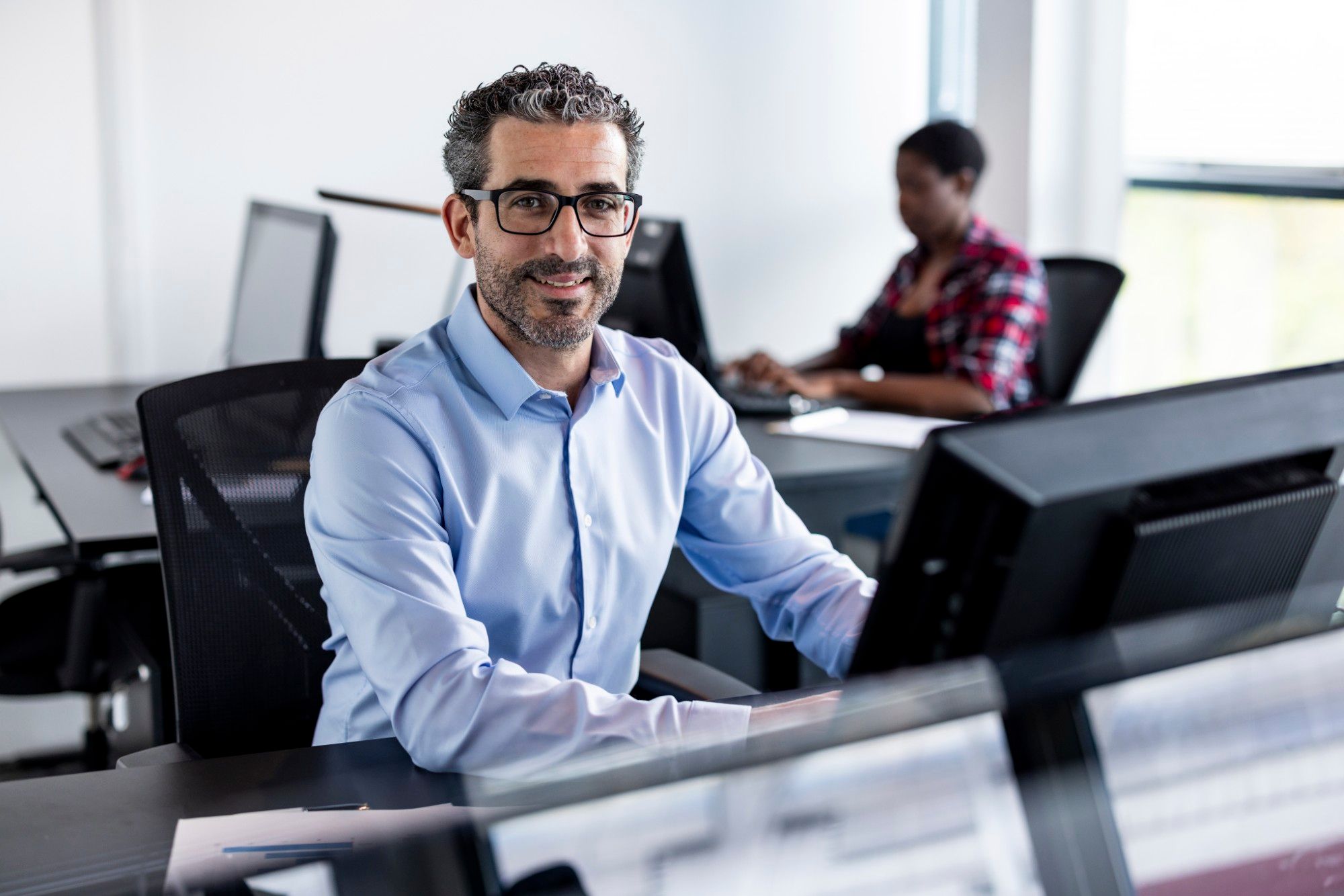 Smiling man with glasses and a beard in a light blue shirt working at a desktop computer in a bright office