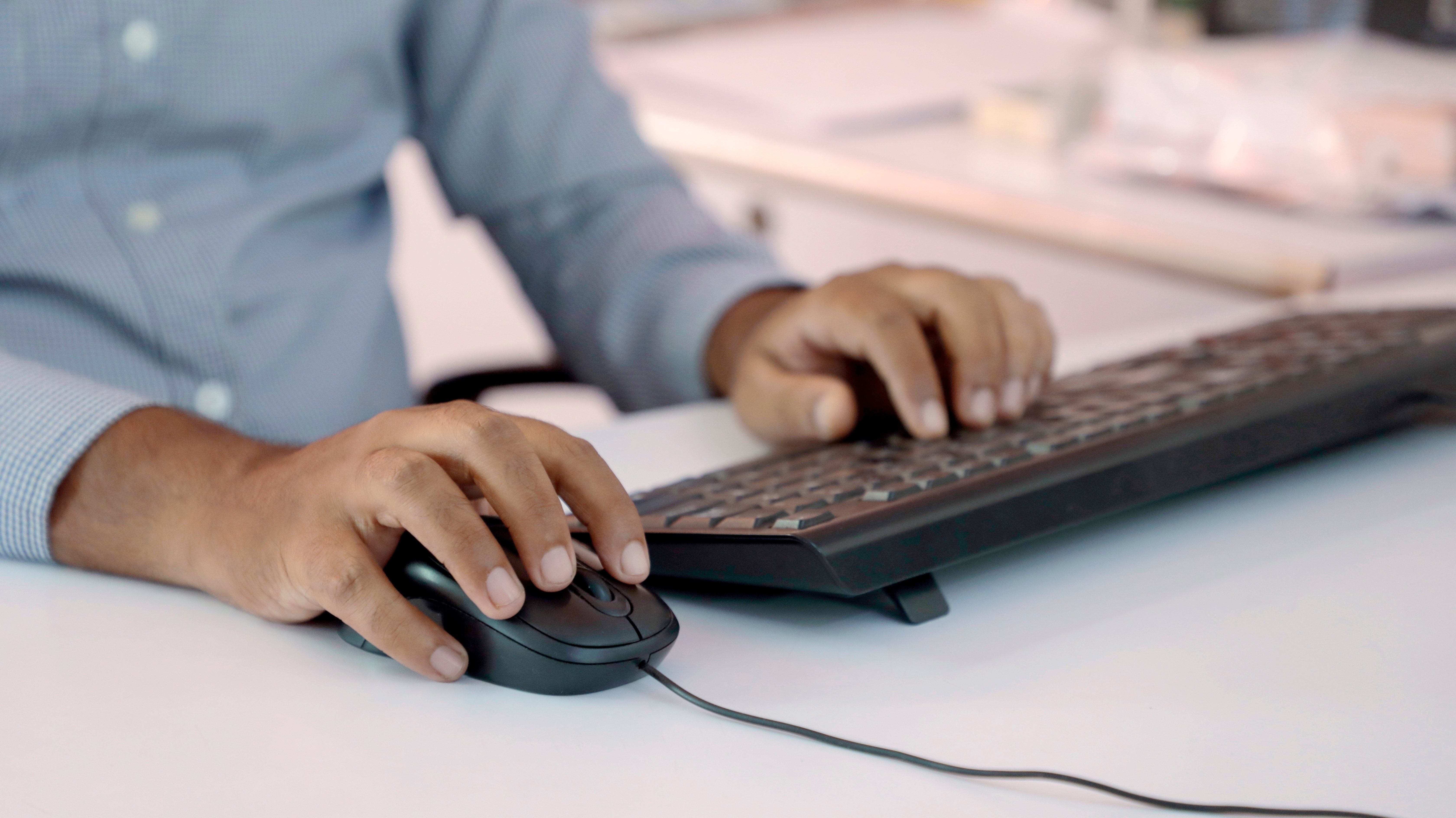 Close-up of a man's hands in a blue shirt using a keyboard and corded mouse at a white office desk