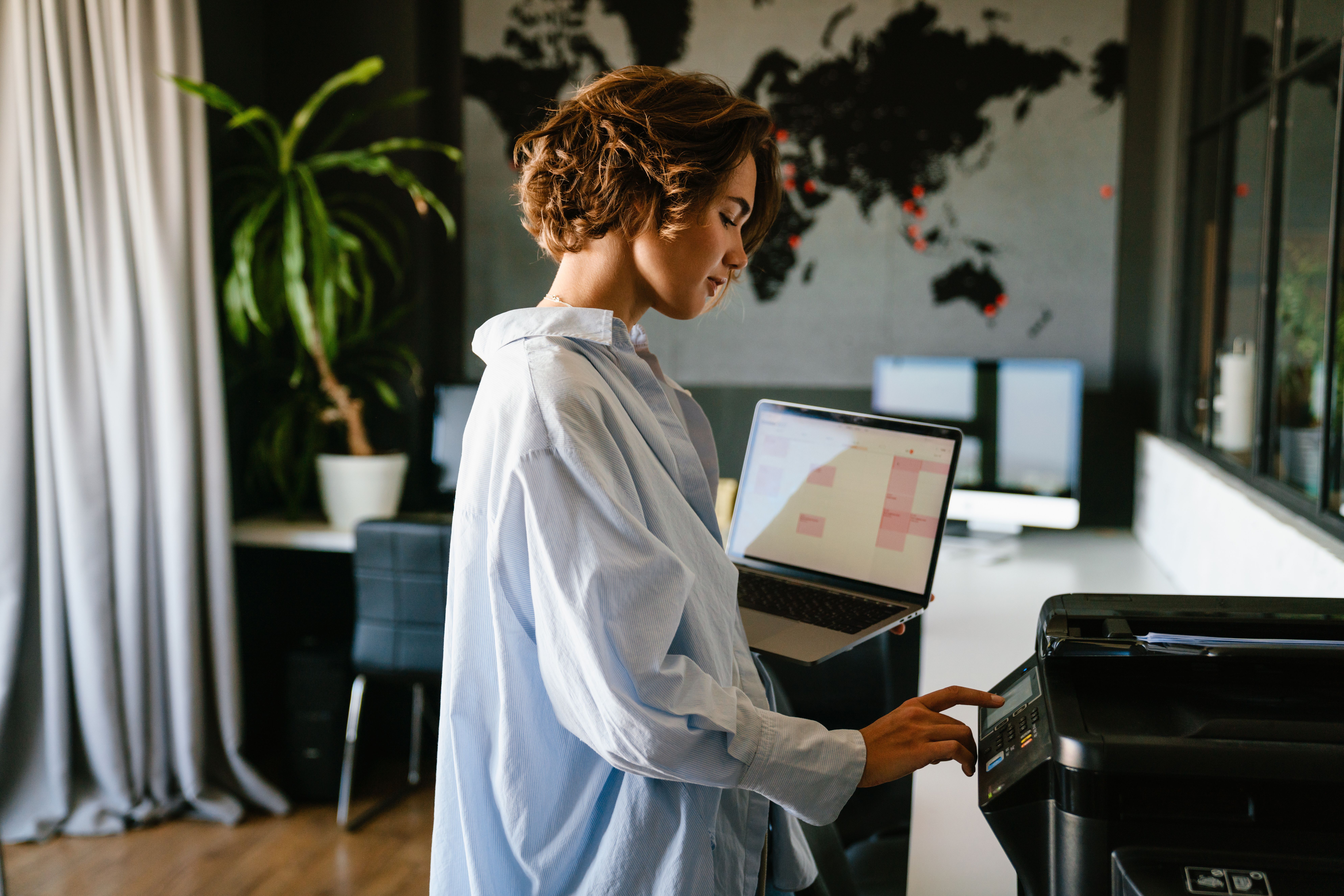 Woman with curly hair using a multifunction printer next to a laptop in a modern office with a world map wall