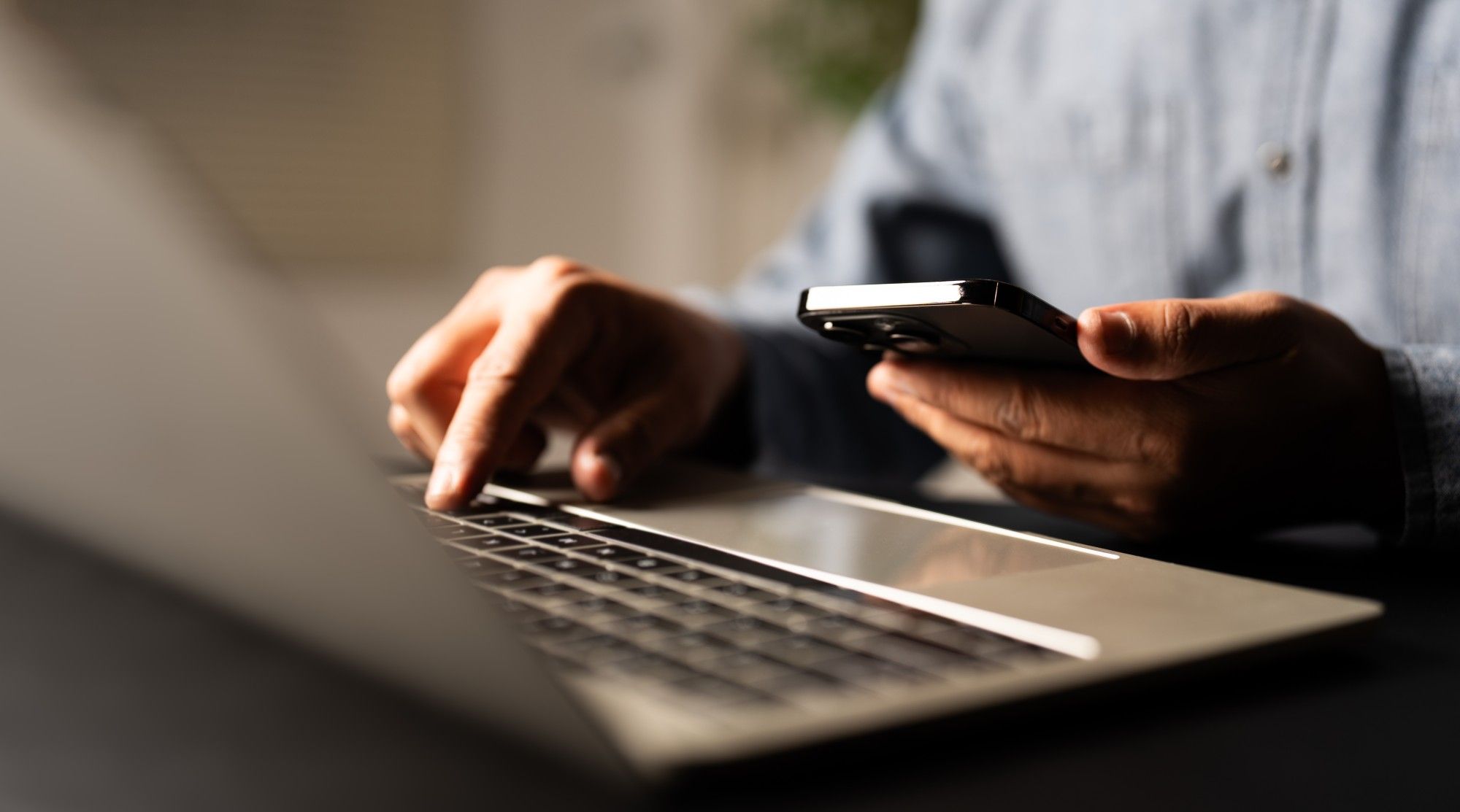 Close-up of a professional's hands typing on a laptop while holding a smartphone in a bright workspace