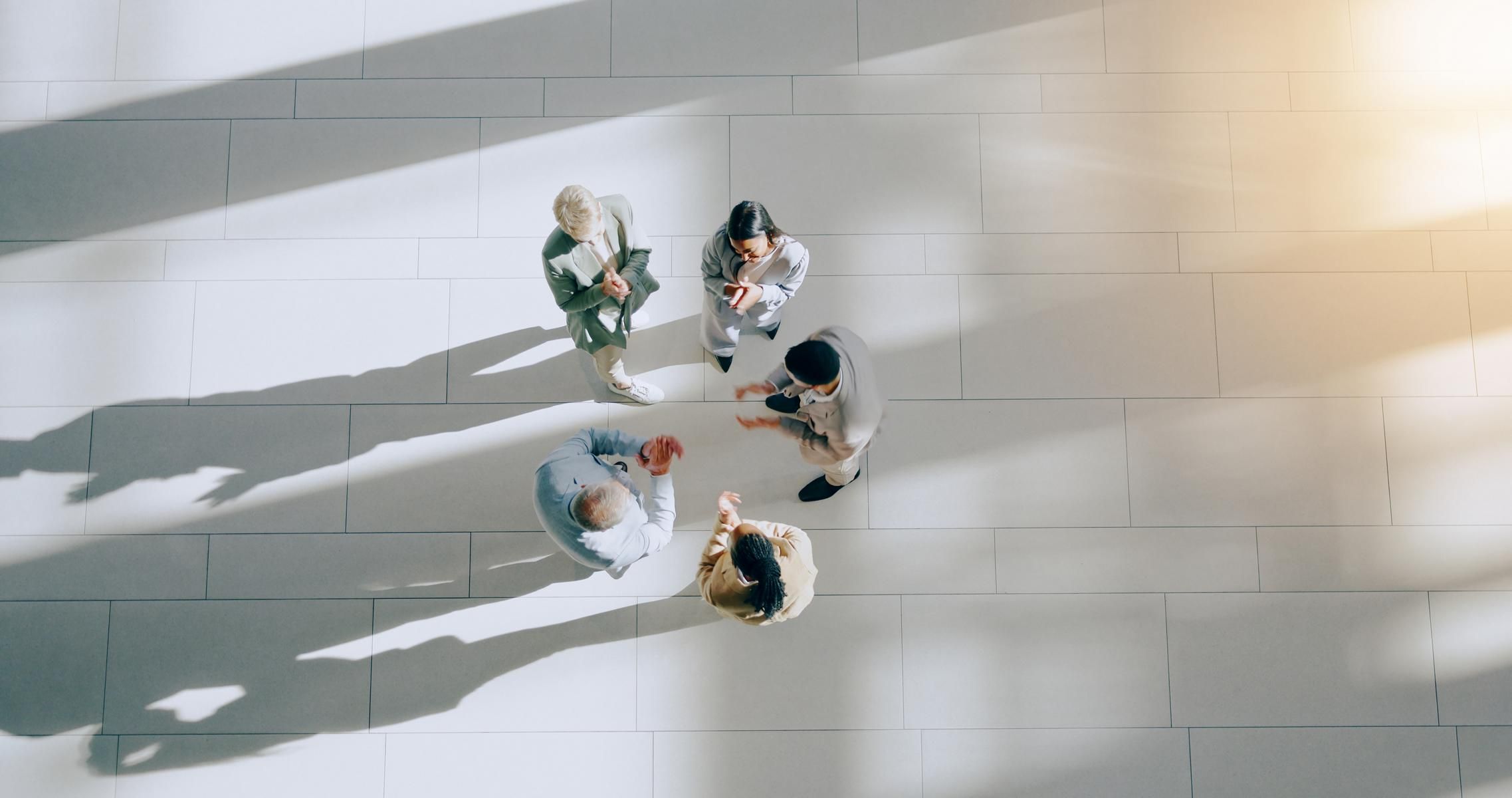 That's image 2 — the couch shot is gone, replaced with this overhead group. Descriptive alt text:  An overhead view of five colleagues standing in a circle and talking on a sunlit tiled floor, their long shadows stretching across a bright modern office