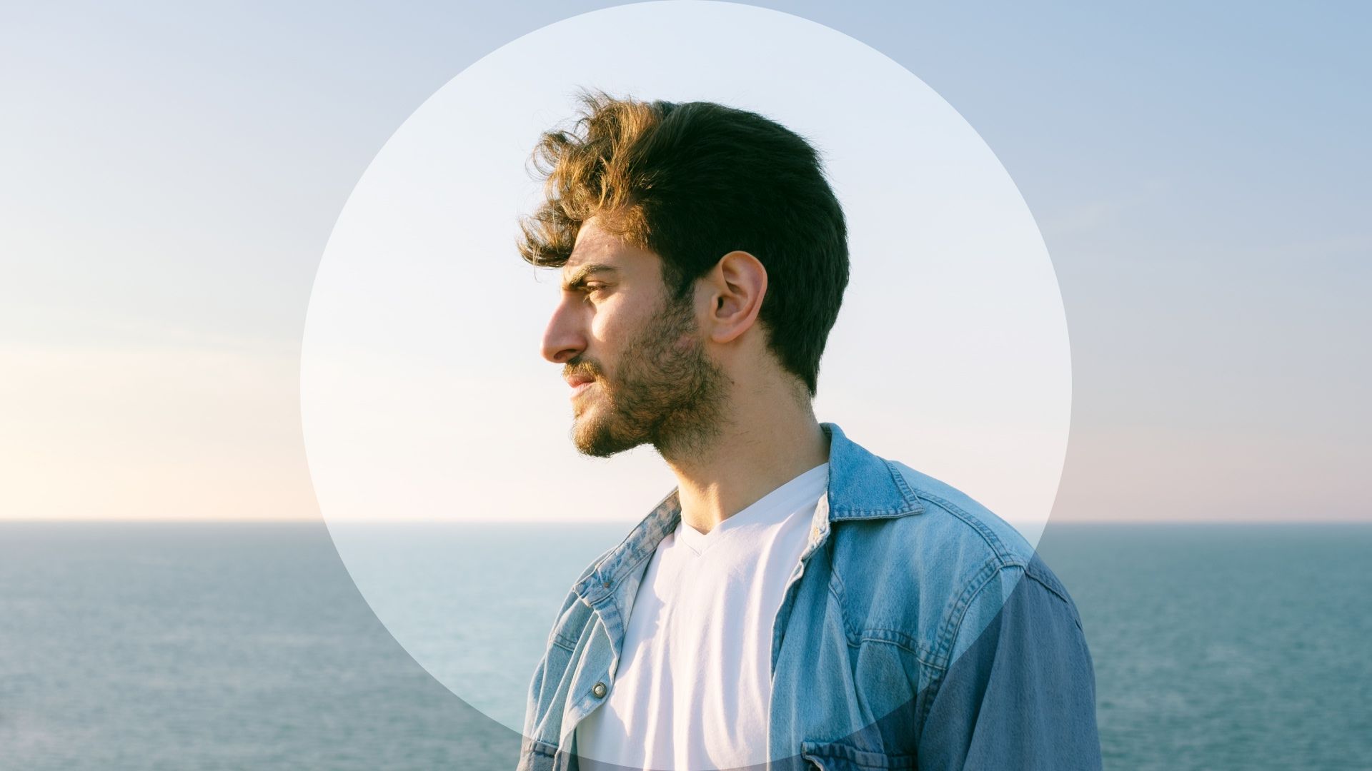 A young bearded man in an open denim shirt stands in profile beside the sea, gazing thoughtfully toward the horizon under a clear sky