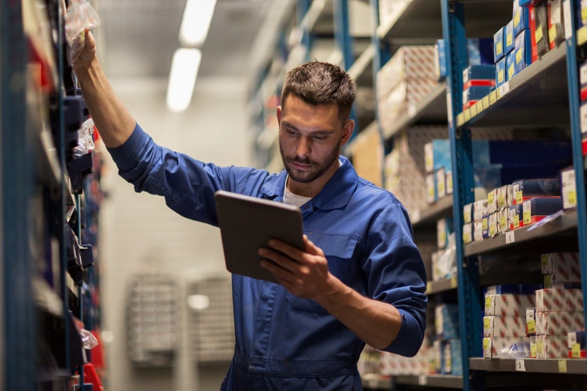 Warehouse worker in blue coveralls checking inventory on a tablet while reaching toward a storage shelf