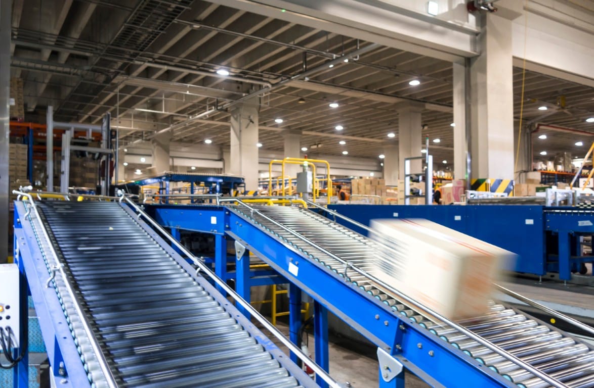 Blue conveyor system in a warehouse with a motion-blurred cardboard box moving along the rollers, surrounded by pallet racking and overhead lighting