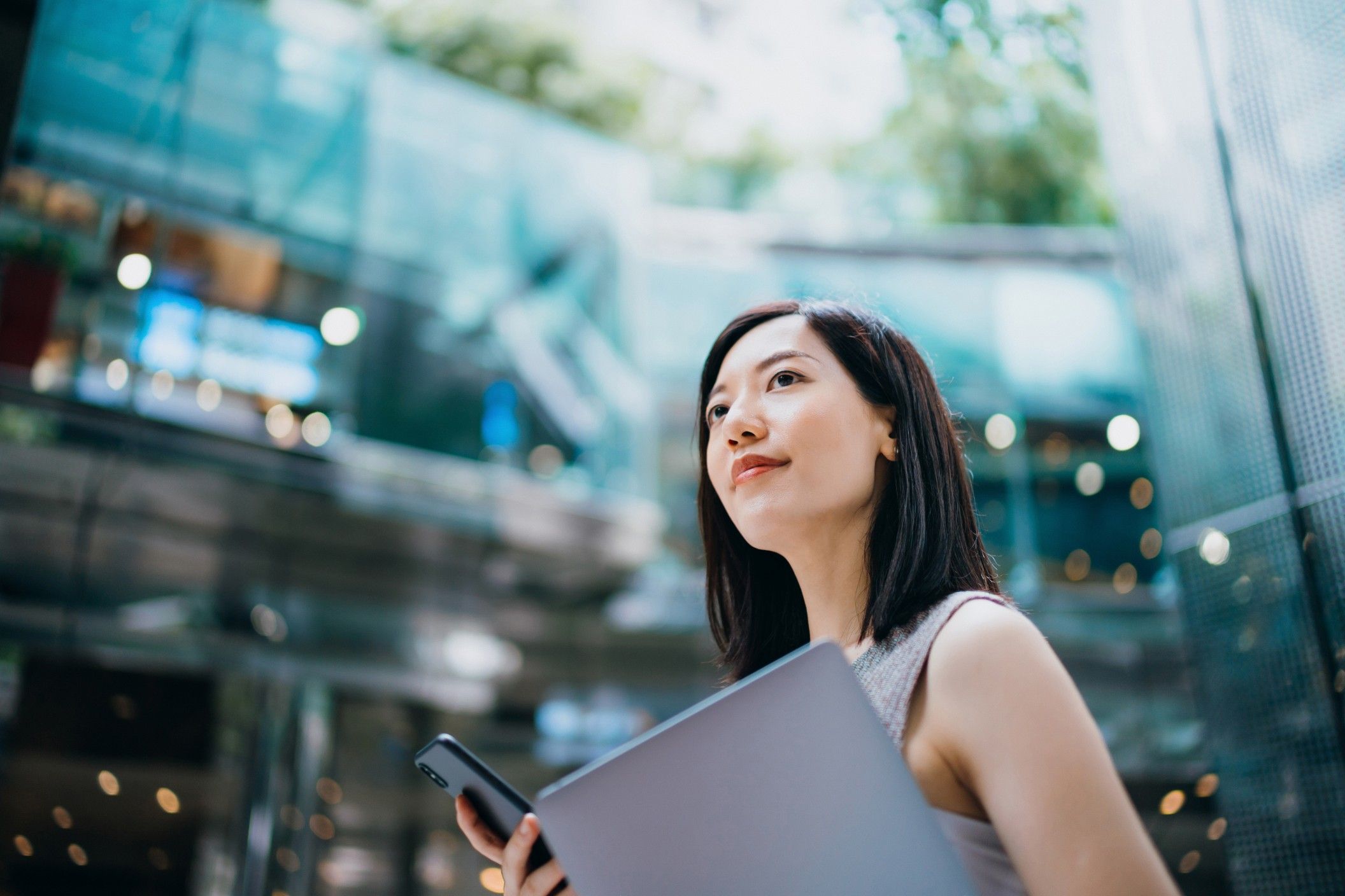 Woman with shoulder-length dark hair carrying a company laptop and smartphone in a modern glass office building