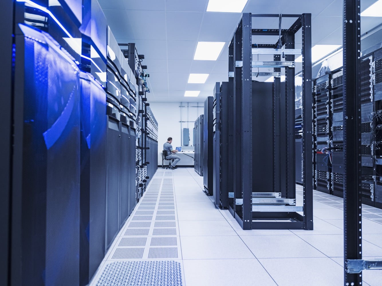 Long aisle of server racks in a data center, with a technician working at a desk at the far end