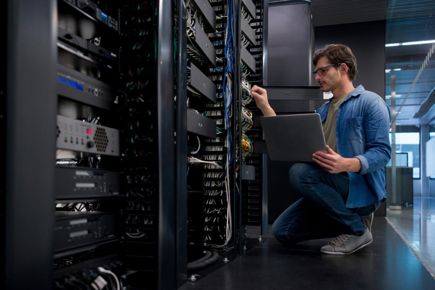 IT engineer kneeling beside open server racks in an enterprise data center, checking hardware with a laptop