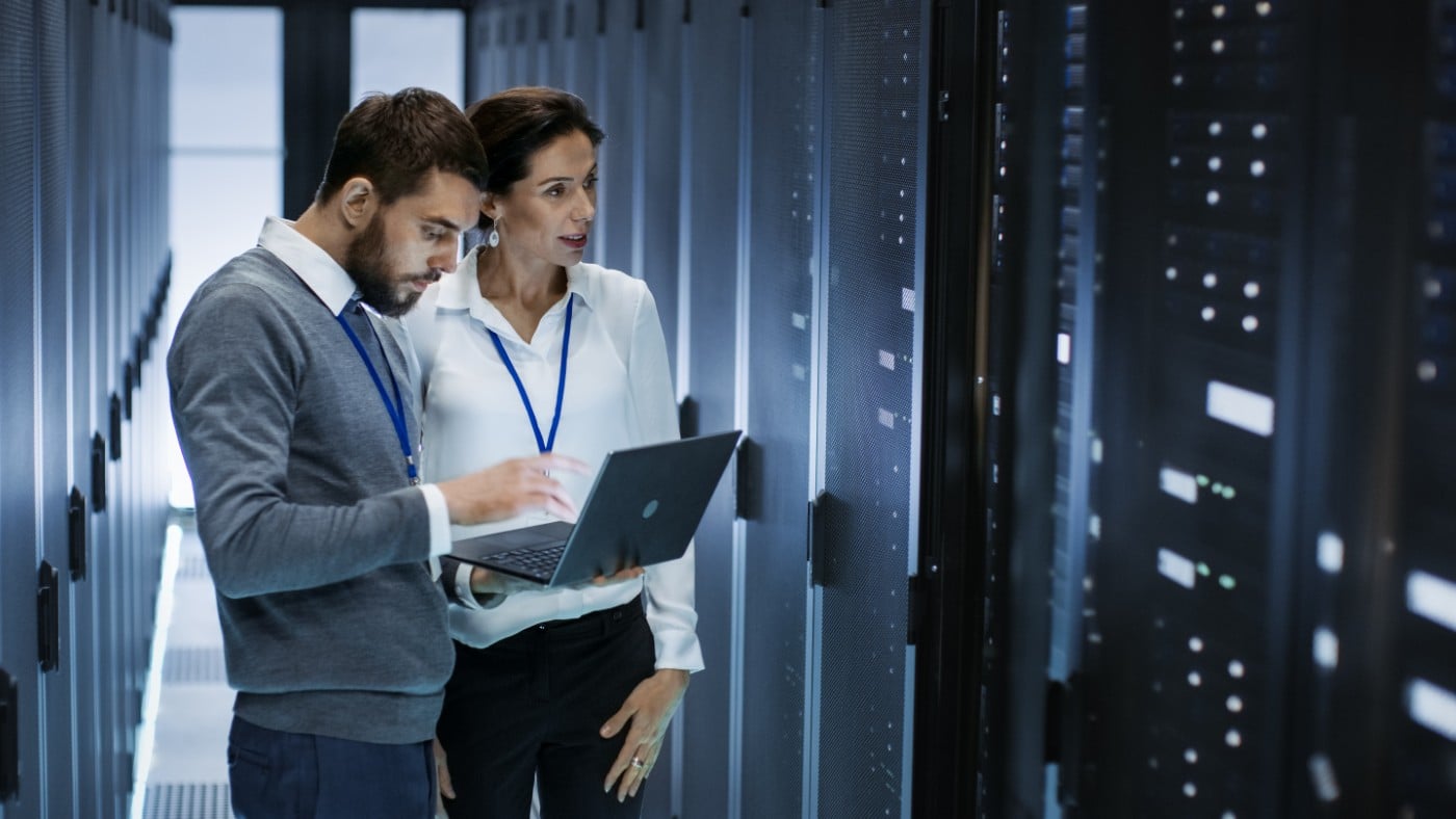 Two IT professionals reviewing AI workload data on a laptop in a data center aisle lined with server racks